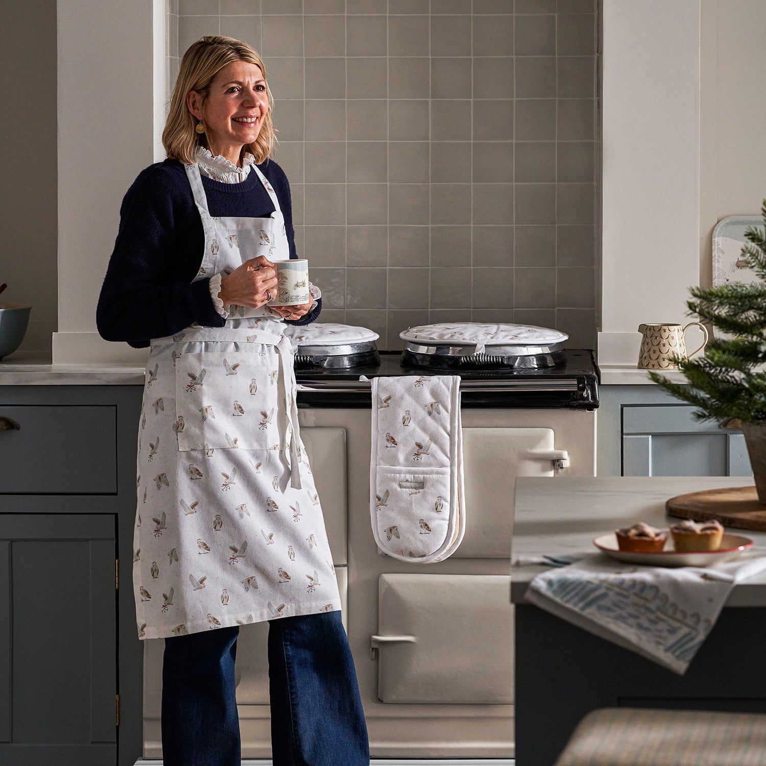 Designer Sophie Allport wearing her Snowy Owl Apron in front of her range cooker and holding a fine bone china mug